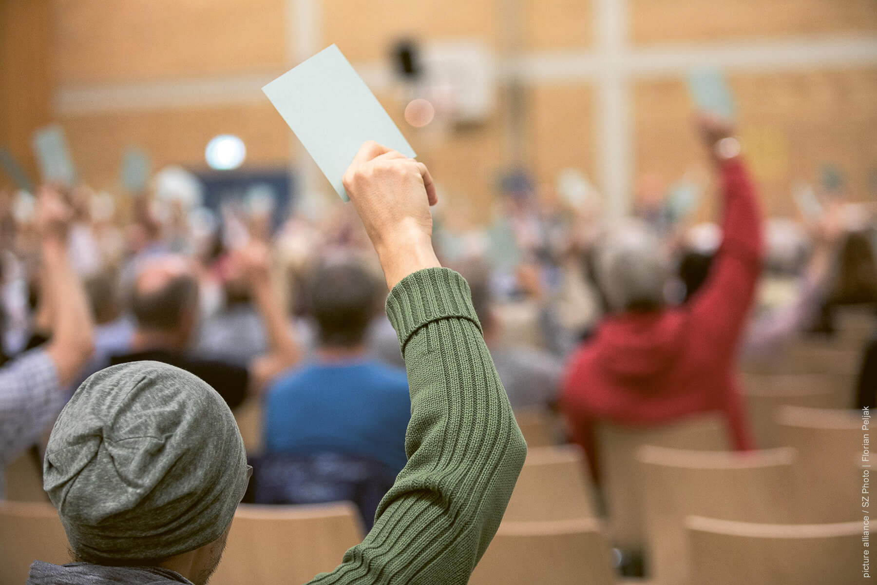 Tag der Demokratie 2021: Sprechblasen Dialog wird als animierte Grafik in ein Foto eingeblendet. Das Foto zeigt eine typische Bürger*innen-Veranstaltung mit einer demokratischen Abstimmung. Die abstimmenden Personen sind unscharf dargestellt.